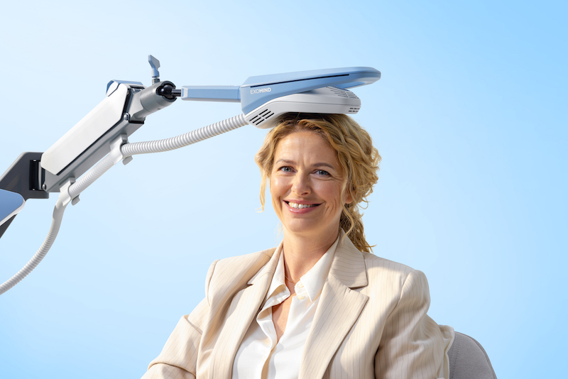 A woman sits down and smiles with an Exomind coil positioned over her scalp as a PTSD treatment in River North Chicago.